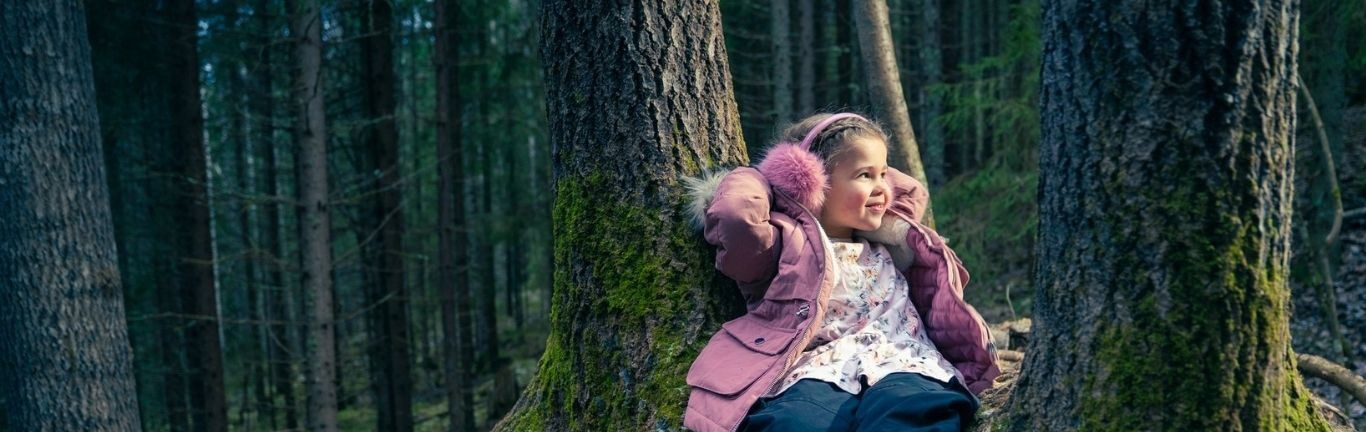 Metsäkuva, jossa tyttölapsi istuu maassa puun juurella.