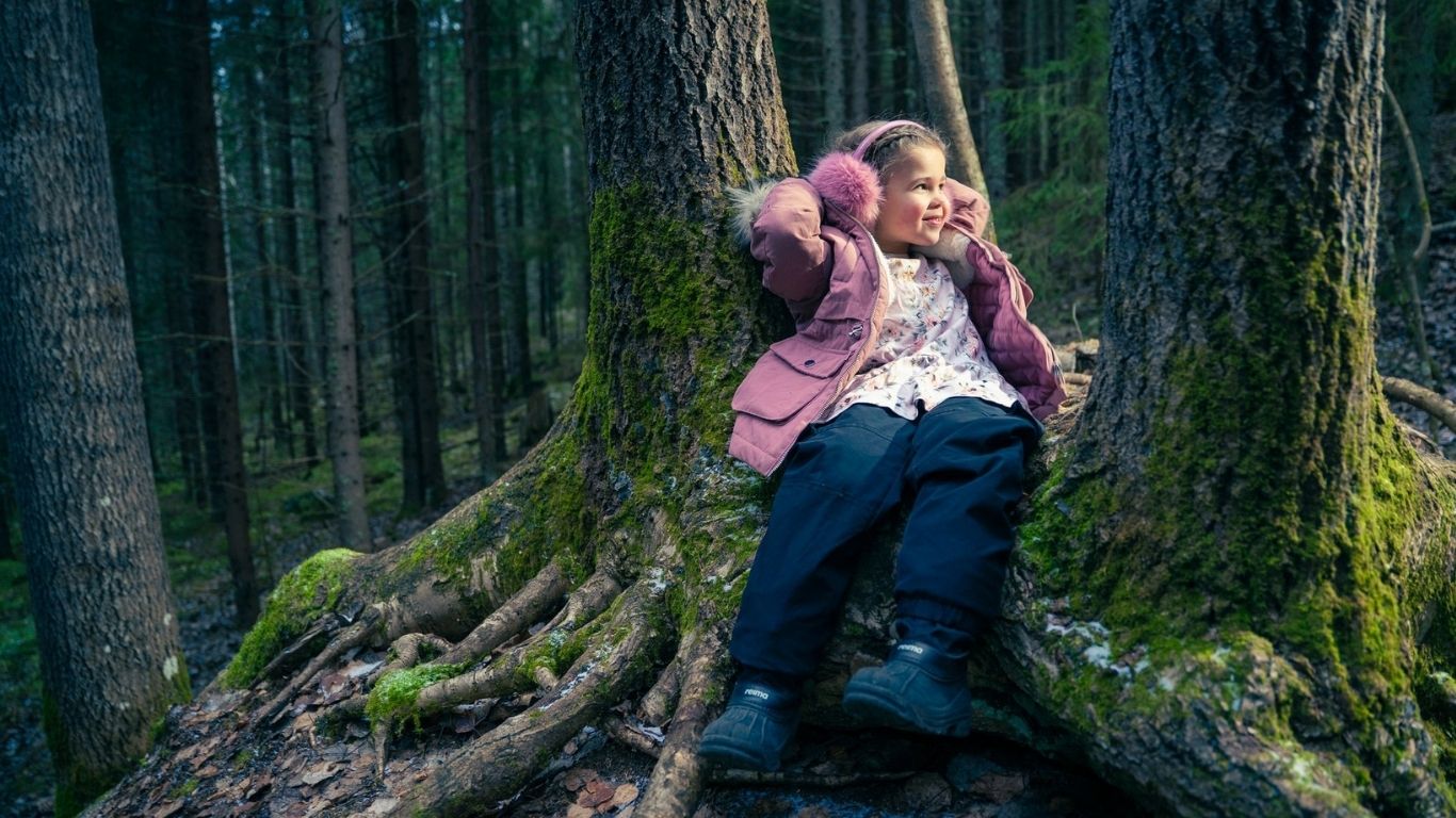Metsäkuva, jossa tyttölapsi istuu maassa puun juurella.